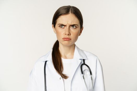Angry and upset young woman doctor, female healthcare worker sulking, frowning disappointed, dislike smth, standing over white background.