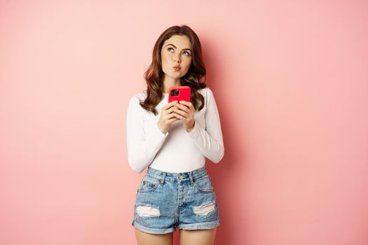 Online shopping. Glamour girl using smartphone to order in app internet, holding mobile phone, thinking, and smiling, looking up thoughtful, standing over pink background.