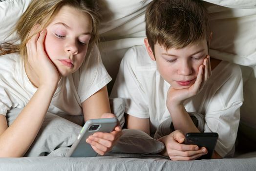 children play games on a smartphone at night under a blanket, rewrites with friends on social networks. social media addiction.
