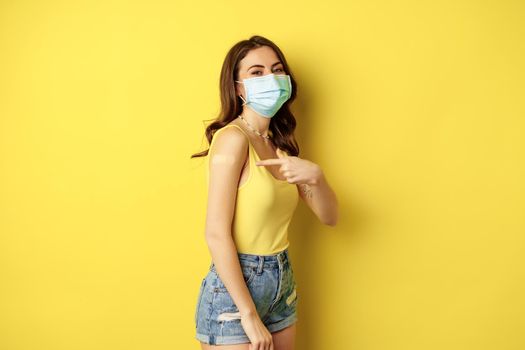 Covid-19 vaccination. Beautiful young woman in face medical mask got vaccinated during pandemic, standing over yellow background.