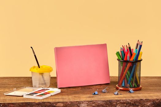 Pink canvas, watercolor set, paintbrush, binder clips, felt-tip pens and multicolored pencils in stand on wooden desktop against beige background. Close up, copy space for your text or images. Mock up