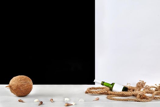 Ripe coconut, colorful seashells, green glass bottle with copy space and rolled sheet of paper in it, thick rope on surface against black and white studio background. Travelling and tourism. Mock up