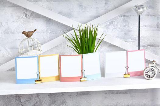 Colorful reminders, blank pieces of paper clamped by binder clips on white shelf, green plant and figurine of bird sitting on decorative cage, candle in silvery candlestick. Close up, copy space