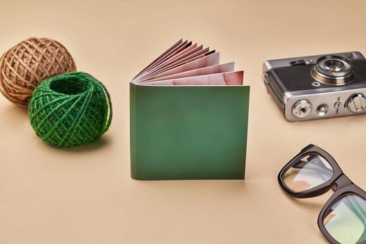 Photo book with green cover, old vintage camera, glasses and two colorful skeins of jute twine or thread on beige studio background. Close up, copy space for your text or images. Mock up, side view