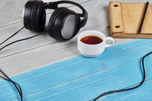 Black headphones with cord, brown notebook in leather cover, pencil and cup of tea on blue and white wooden tabletop. Modern technologies. Close up, copy space for your text or images