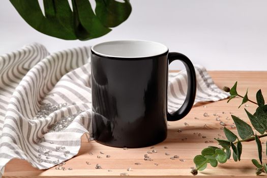 Black porcelain mug for coffee or tea on wooden desktop next to striped tablecloth, scattered crystals and green twigs against white studio background. Mock up, branding area. Close up, copy space