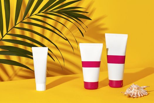 One white and two colorful cosmetic tubes with no logo next to a shell and green palm twig on yellow studio background. Packaging, containers. Advertising, branding area. Close up, copy space, mock up