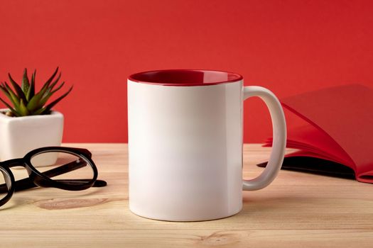 White porcelain mug for coffee or tea on wooden desktop next to notepad, glasses and green succulent in pot against red studio background. Mock up, branding area. Close up, copy space