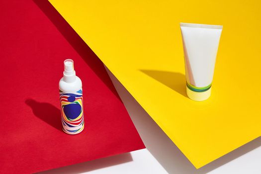 Colorful cosmetic tube and pump bottle with no logo standing on red and yellow sheets of paper above white surface. Packaging, containers. Advertising, branding area. Close up, copy space, mock up