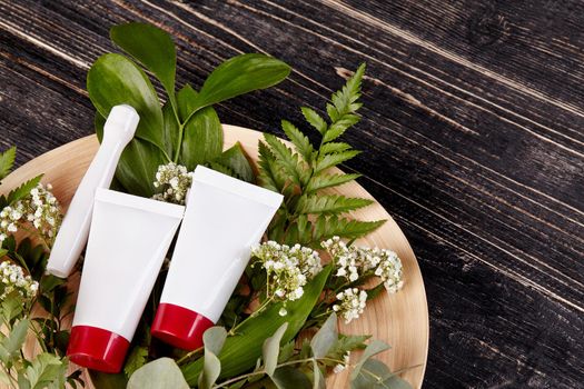 Two white cosmetic tubes with red caps and roller for face on bouquet of flowers lying on plate against wooden background. Packaging, advertising. Close up, copy space. Top view, mock up