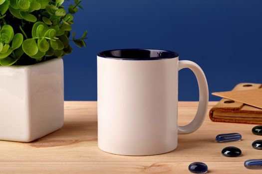 White ceramic mug on wooden desktop next to scattered decorative glass stones, green plant in pot and brown notepad against blue studio background. Mock up, branding area. Close up, copy space