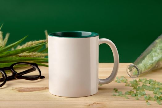 White ceramic mug, coarse sea salt scattered from the bottle on wooden desktop, glasses and blooming spikelets against green studio background. Mock up, branding area. Close up, copy space