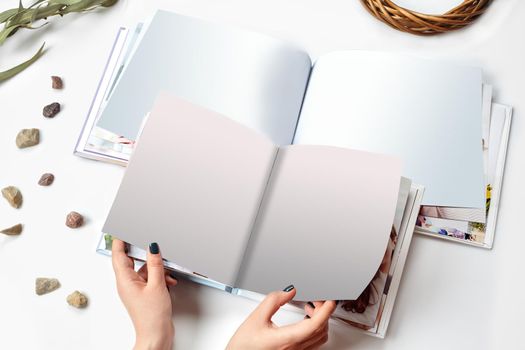 Hands of a female who holding two open photo books with hardback and blank pages, green twig and small stones isolated on white studio background. Close up, copy space. Mock up, top view, flat lay