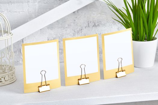 Three small blank pieces of paper clamped by binder clips on white shelf next to green plant in pot and decorative birds cage. Close up, copy space for your text or images. Mock up, interior