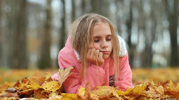 A little girl indifferently lies on the autumn foliage in a city park