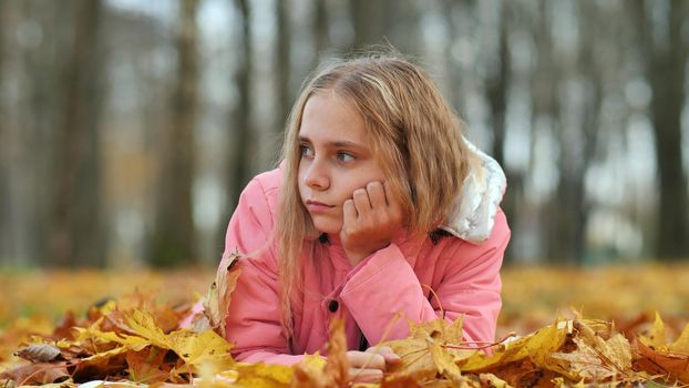 A little girl indifferently lies on the autumn foliage in a city park. She is very sad