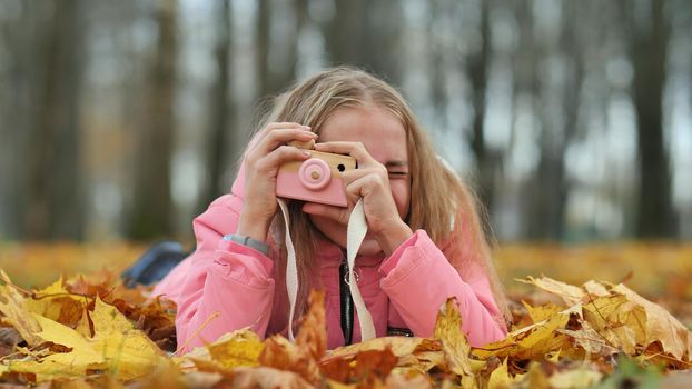 A teenage girl with a toy camera takes pictures of lying in the autumn foliage in the park