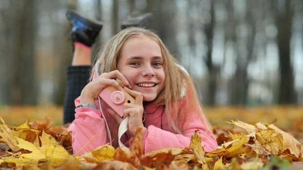 A teenage girl with a toy camera takes pictures of lying in the autumn foliage in the park