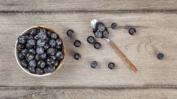 Table top view on bowl of blueberries, some of them spilled next to spoon on gray wood desk.