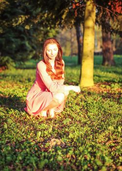 Young woman in pink dress crouching in the park, during evening sunset light.