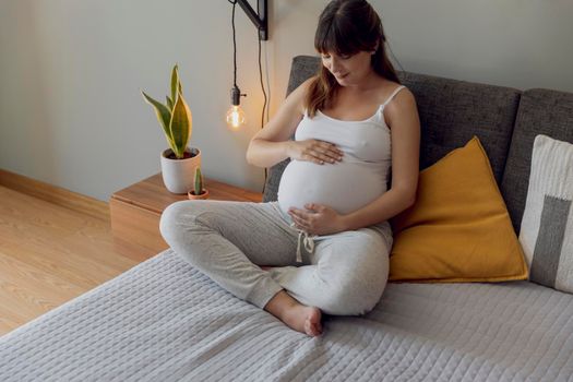 Happy pregnant woman sitting on bed and touching her belly at home