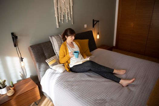 Beautiful woman on bed with a cup of coffee and reading a book