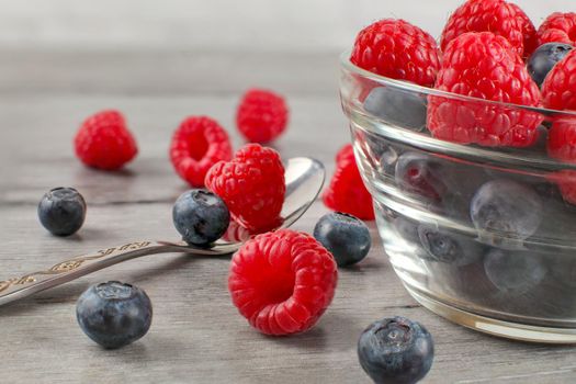 Small glass bowl full of blueberries and raspberries mix, some spilled on gray wood desk with silver spoon.