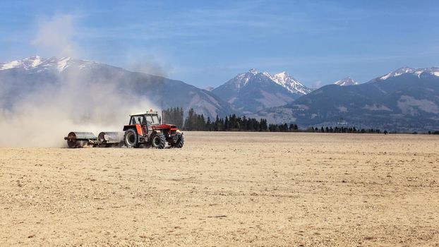 Tractor pulling metal rollers over dry field with mountains in background. Spring soil preparation.