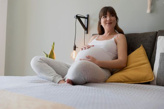 Happy pregnant woman sitting on bed and touching her belly at home