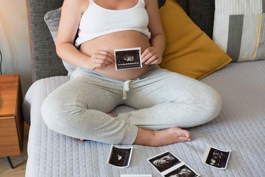Pregnant woman holding the picture of the ultrasound on her belly in bed at home. Prenatal health care concept.