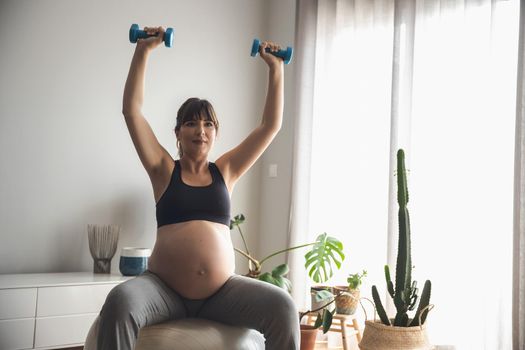 Pregnant woman working out with dumbbells doing strength exercises on a fitball at home. Keeping in good shape while waiting for baby