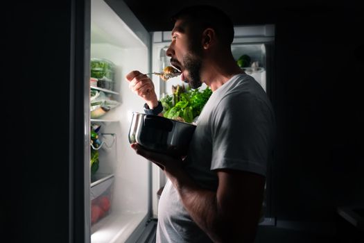 Young hungry man eating food at night from open fridge. Midnight snack and food addiction. High quality photo