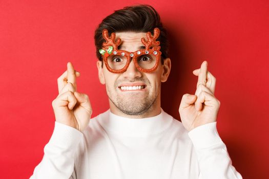 Concept of winter holidays, christmas and celebration. Close-up of nervous man in party glasses, crossing fingers for good luck and pleading, making a wish, standing over red background.