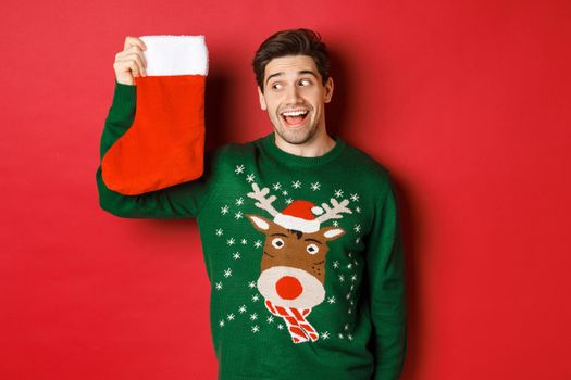 Image of surprised and amused man in green sweater, looking at christmas stocking with presents and smiling, standing over red background.