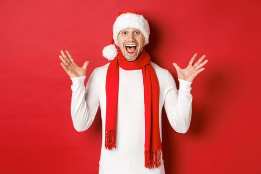 Portrait of happy and amazed handsome man, celebrating new year, wishing merry christmas, wearing santa hat and scarf, telling big news, standing against red background.