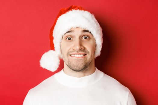 Close-up of handsome guy in santa hat, biting lip and looking with temptation at something he wants, standing over red background.