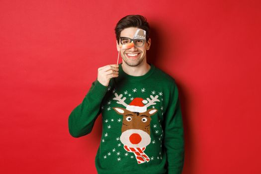 Funny man in christmas sweater and party mask, celebrating winter holidays, smiling happy, standing over red background.