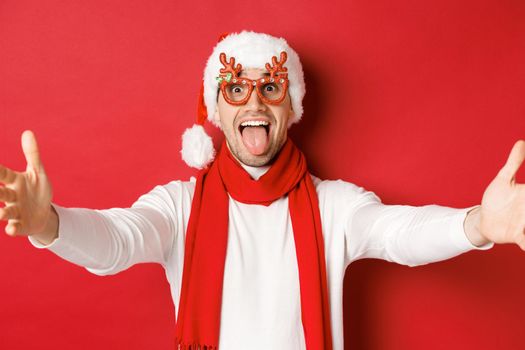 Concept of christmas, winter holidays and celebration. Close-up of happy smiling man in party glasses and santa hat, spread hands sideways for hug, greeting guests, red background.