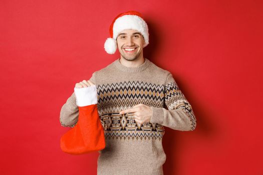 Concept of winter holidays, new year and celebration. Handsome smiling man prepared gifts for kids, pointing at christmas stocking bag, standing over red background.