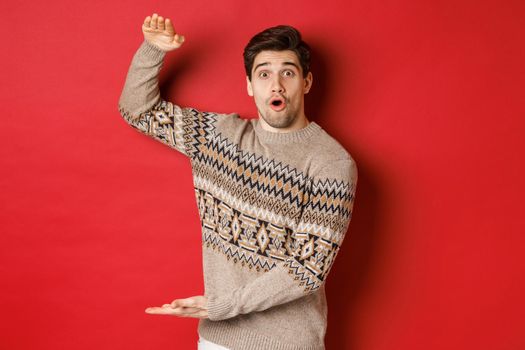 Image of attractive man in christmas sweater, shaping big new year gift, showing something large and amazing, standing over red background.