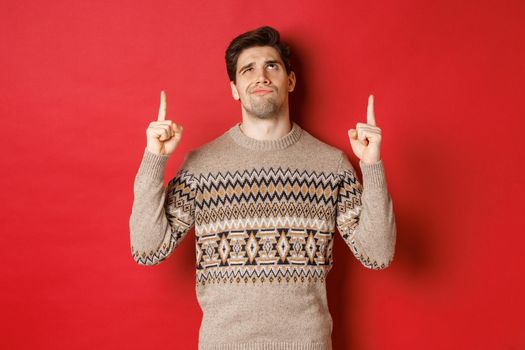 Image of disappointed and skeptical handsome man, wearing christmas sweater, smirk and frowning as looking at something bad, pointing fingers up, standing over red background.