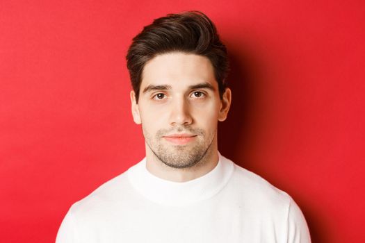 Close-up of handsome brunette guy in white sweater, smiling at camera, standing over red background.