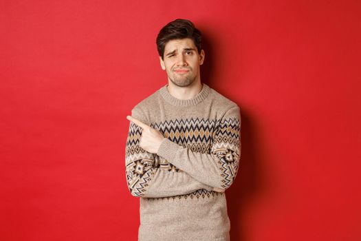 Image of skeptical and displeased man in christmas sweater, pointing at upper left corner unamused, showing bad xmas gift, standing over red background.