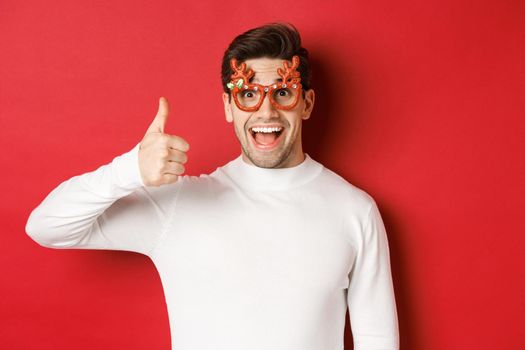 Close-up of handsome young man in party glasses and white sweater, showing thumb-up in approval, enjoying new year holidays, standing over red background.