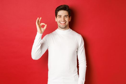 Concept of winter holidays, christmas and lifestyle. Happy handsome man in white sweater showing okay sign, smiling pleased, recommending shop, standing over red background.