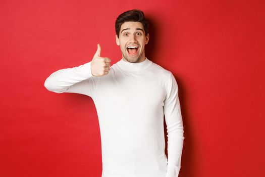 Concept of winter holidays, christmas and lifestyle. Excited good-looking man in white sweater, like good product, showing thumb-up in approval and smiling amazed, red background.