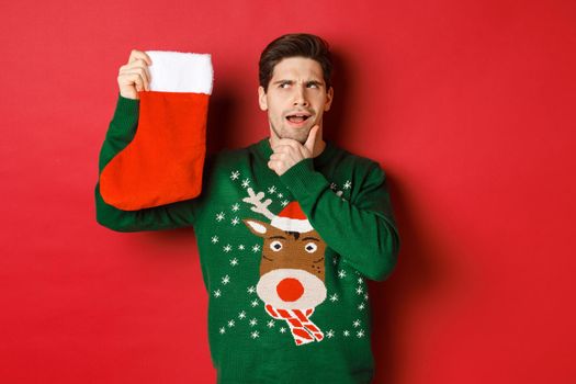 Portrait of thoughtful handsome man in green sweater, holding christmas stocking and thinking, guessing what present is inside, standing over red background.