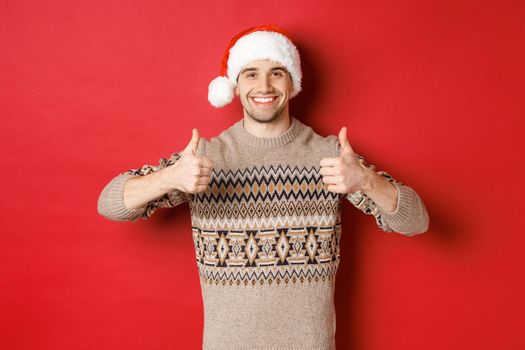 Image of cheerful attractive man in winter sweater and santa hat, showing thumbs-up, celebrating christmas and looking happy, like something, standing over red background.