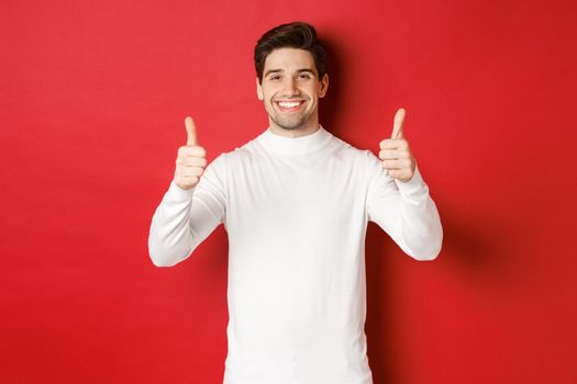 Concept of winter holidays, christmas and lifestyle. Portrait of satisfied handsome man in white sweater showing thumbs-up, praise something good, like it, standing over red background.