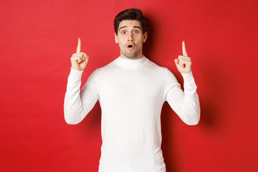 Concept of winter holidays. Good-looking young man with bristle, wearing white sweater, showing christmas advertisement on copy space, pointing fingers up and looking surprised, red background.